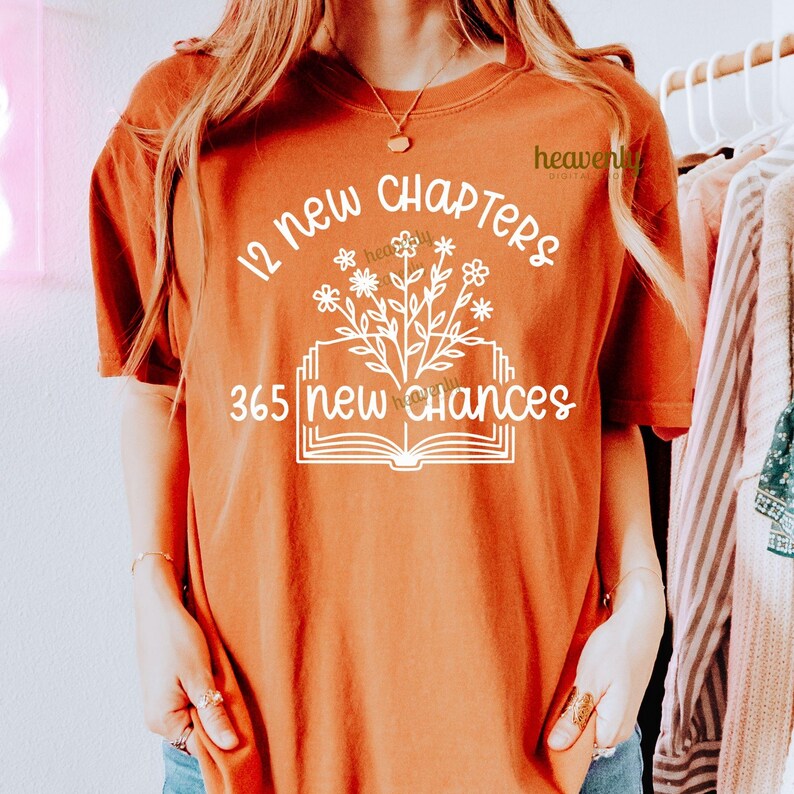 May include: A rust-colored t-shirt with a white graphic design that reads "12 new chapters 365 new chances". The graphic features a book with a flower growing out of it.