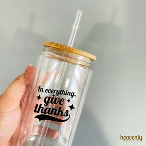 May include: A clear glass tumbler with a bamboo lid and a straw. The tumbler has a black vinyl decal that says "In everything, give thanks" with a star on each side of the word "give".
