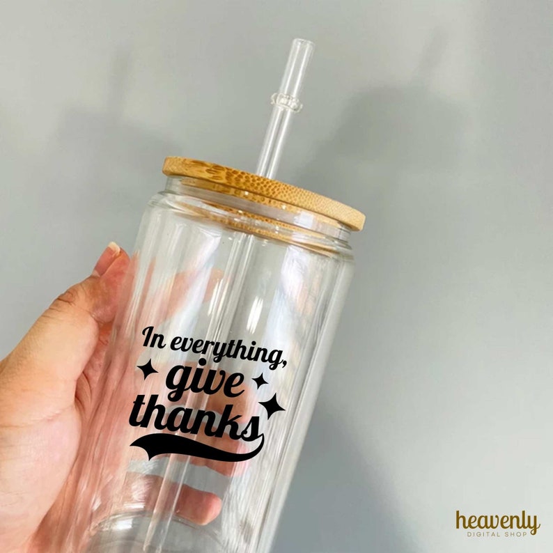 May include: A clear glass tumbler with a bamboo lid and a straw. The tumbler has a black vinyl decal that says "In everything, give thanks" with a star on each side of the word "give".