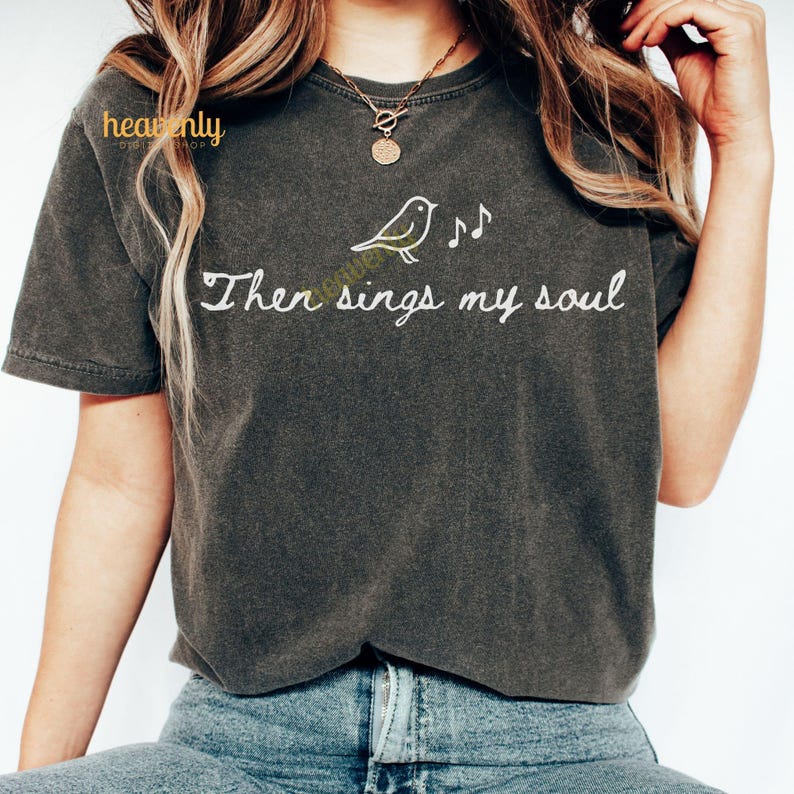 May include: Dark gray t-shirt with the text "Then sings my soul" in white script. A small bird and musical notes are printed above the text. The shirt is a crew neck and has short sleeves.
