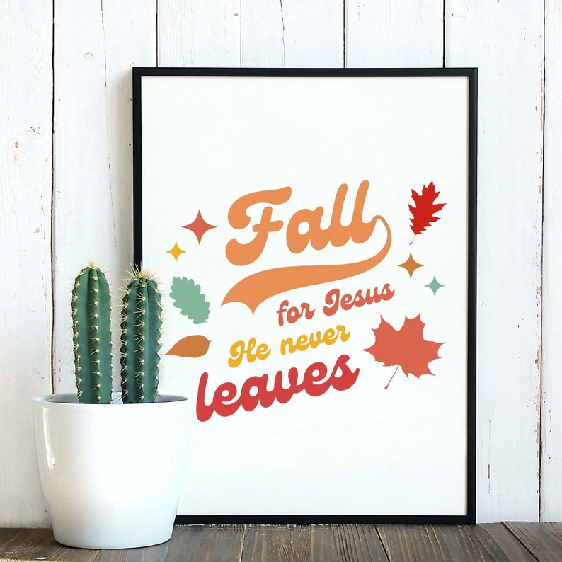 Op de afbeelding: Een wit ingelijst print met de tekst "Fall for Jesus He never leaves" in een kleurrijk retro-lettertype. De print bevat gestileerde herfstbladeren in oranje, rood en geel.