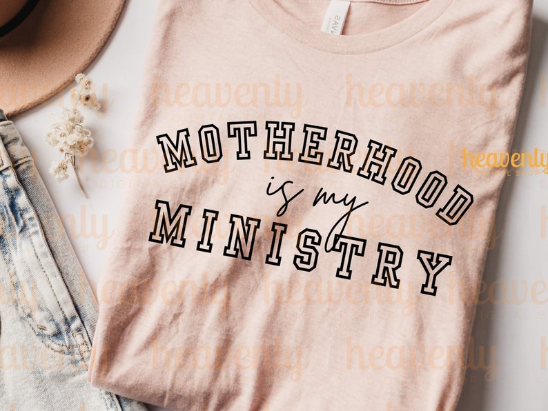 Pu&ograve; includere: Una maglietta rosa con il testo "MOTHERHOOD is my MINISTRY" in lettere nere con contorno.