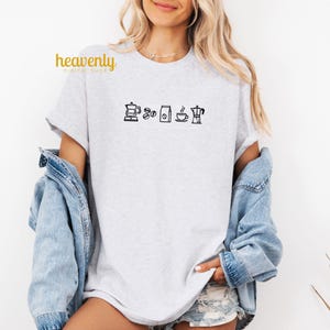 May include: A gray t-shirt with a black graphic of coffee icons, including a coffee pot, coffee beans, a bag of coffee, a cup of coffee, and a coffee maker.