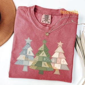 May include: A red Comfort Colors t-shirt featuring three Christmas trees with star toppers. The trees have a patchwork design in shades of blue, green, and pink. The shirt has the text "heavenly digital shop" and a silver necklace.