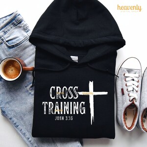 Może przedstawiać: Czarna bluza z kapturem z białym krzyżem i tekstem "CROSS TRAINING JOHN 3:16" w stylu vintage.
