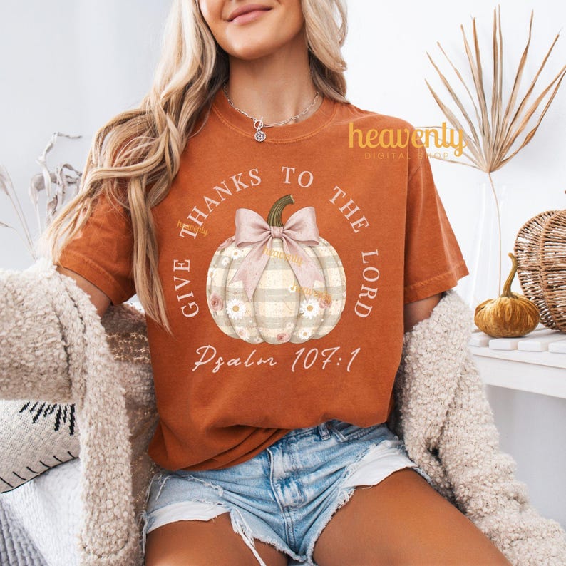 Puede incluir: Camiseta naranja con un dise&ntilde;o de calabaza y el texto "GIVE THANKS TO THE LORD" y "Psalm 107:1". La calabaza tiene un lazo rosa y detalles florales. Esta camiseta es parte de una colecci&oacute;n con tem&aacute;tica de oto&ntilde;o.