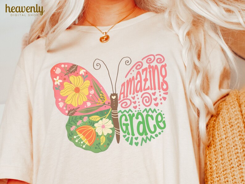 Puede incluir: Una camiseta blanca con un dise&ntilde;o de mariposa colorida. La mariposa tiene flores rosas, verdes, amarillas y naranjas en sus alas. El texto "amazing grace" est&aacute; escrito en rosa y verde en las alas de la mariposa.