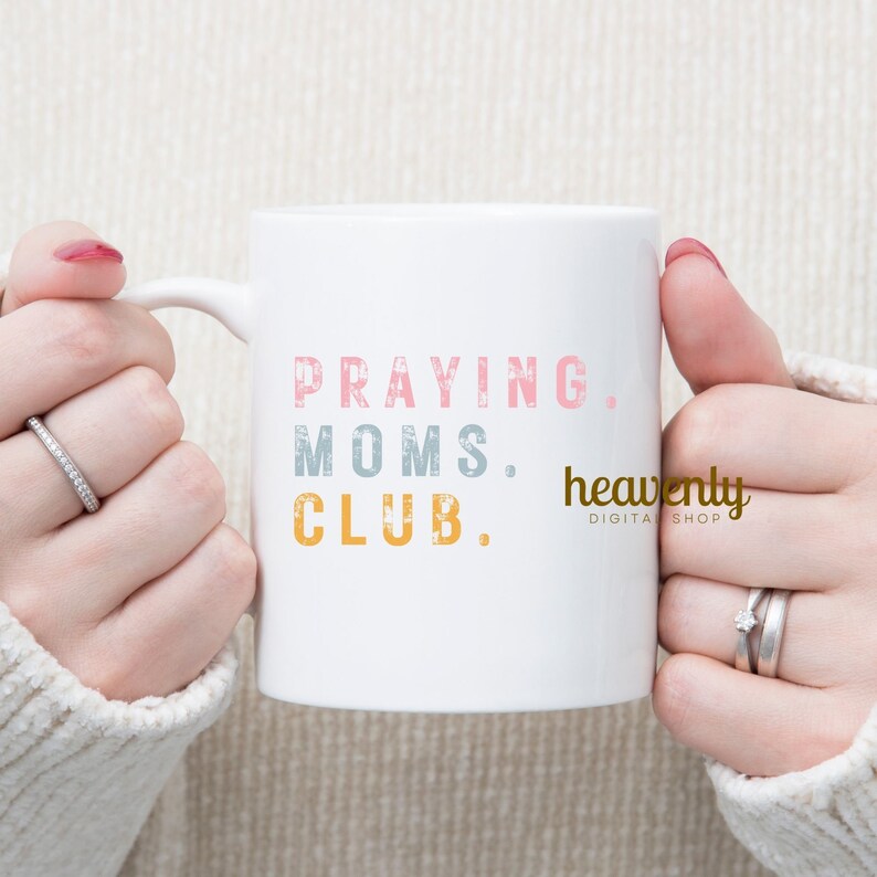 May include: White ceramic mug with the text "Praying Moms Club." in pink, gray, and yellow. The text "heavenly digital shop" is in gray and white.
