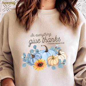 May include: A beige sweatshirt with the text "In everything, give thanks 1 Thessalonians 5:18" printed on the front. The text is surrounded by a watercolour illustration of pumpkins and leaves.