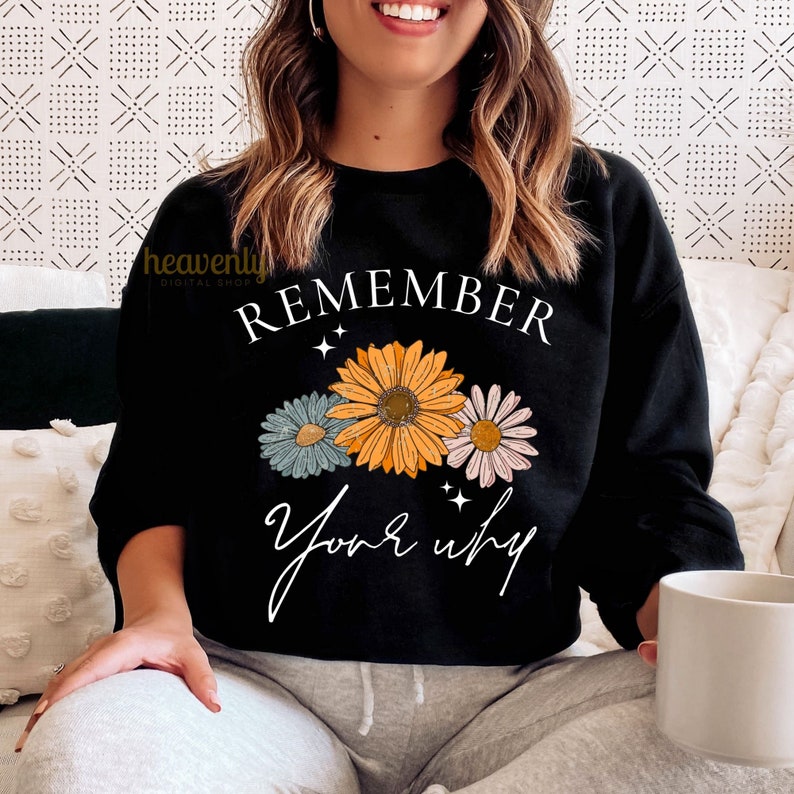 K&ouml;nnte beinhalten: Schwarzer Pullover mit dem Text "REMEMBER your why" in wei&szlig;er Schrift. Der Text ist von drei stilisierten Blumen in Orange-, Rosa- und Blaut&ouml;nen umgeben.