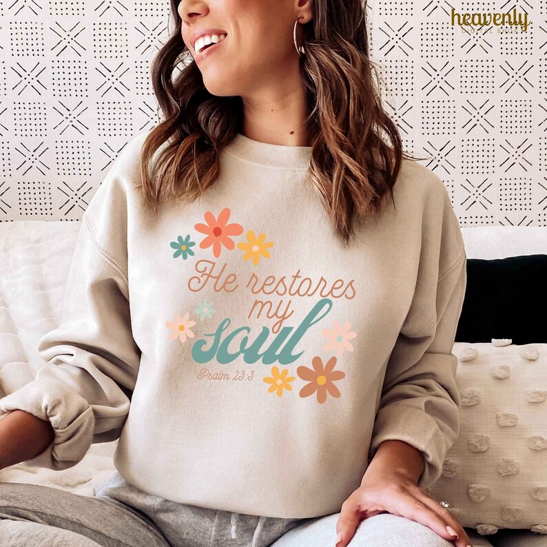 Puede incluir: Una sudadera beige con un dise&ntilde;o floral y el texto "He restores my soul" con la referencia b&iacute;blica "Salmo 23:3".