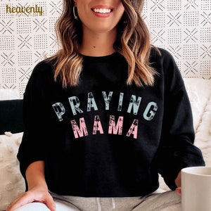May include: Black sweatshirt with the text "Praying Mama" in blue and pink lettering. The letters have a distressed, vintage look.
