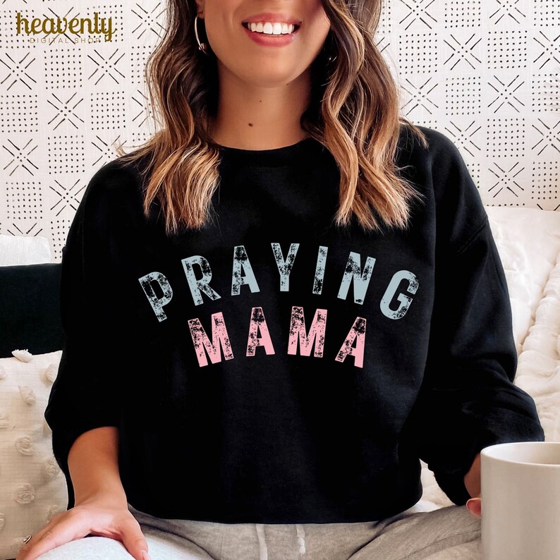 May include: Black sweatshirt with the text "Praying Mama" in blue and pink lettering. The letters have a distressed, vintage look.