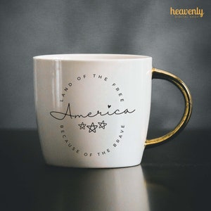 May include: White ceramic mug with a gold handle. The mug has a black circle design with the text "Land of the Free, America, Because of the Brave" and three stars.