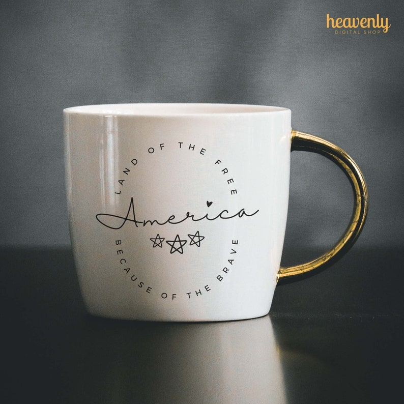 May include: White ceramic mug with a gold handle. The mug has a black circle design with the text "Land of the Free, America, Because of the Brave" and three stars.