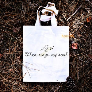 May include: White tote bag with cream-colored handles, featuring a black illustration of a bird with musical notes and the text "Then sings my soul." The bag is set on a bed of brown pine needles.