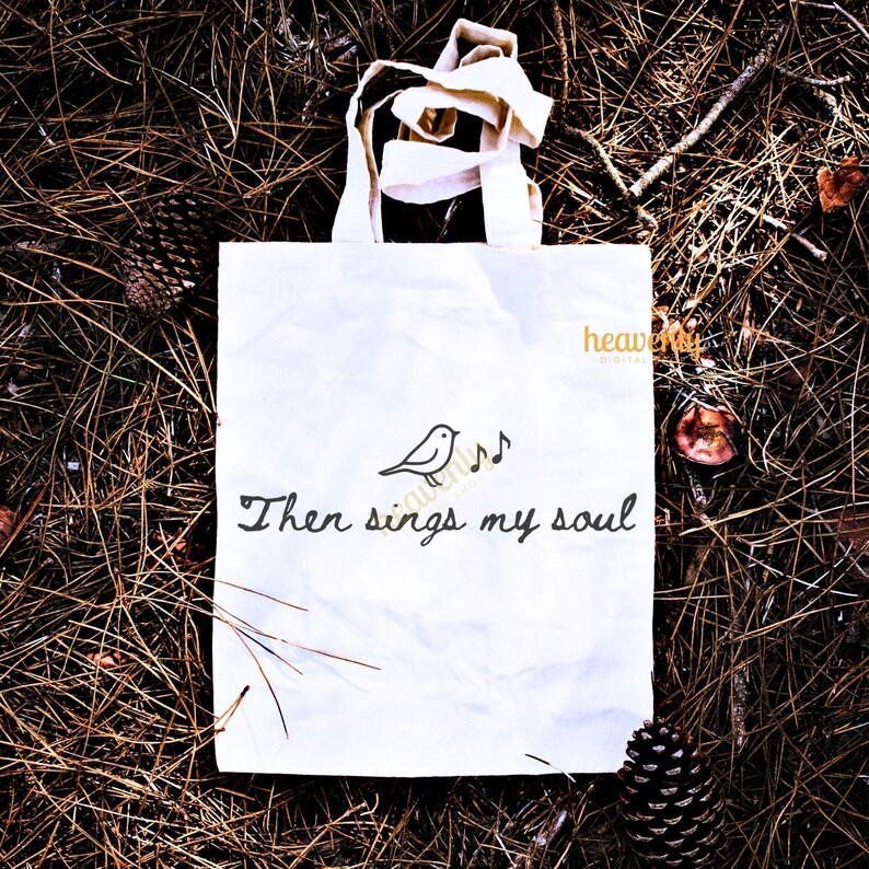 May include: White tote bag with cream-colored handles, featuring a black illustration of a bird with musical notes and the text "Then sings my soul." The bag is set on a bed of brown pine needles.
