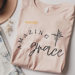 Pu&ograve; includere: Una maglietta rosa chiaro con le parole "Amazing Grace" in corsivo nero. Una croce nera con accenti floreali &egrave; sul lato destro. La maglietta ha anche il testo "heavenly digital shop" in giallo.