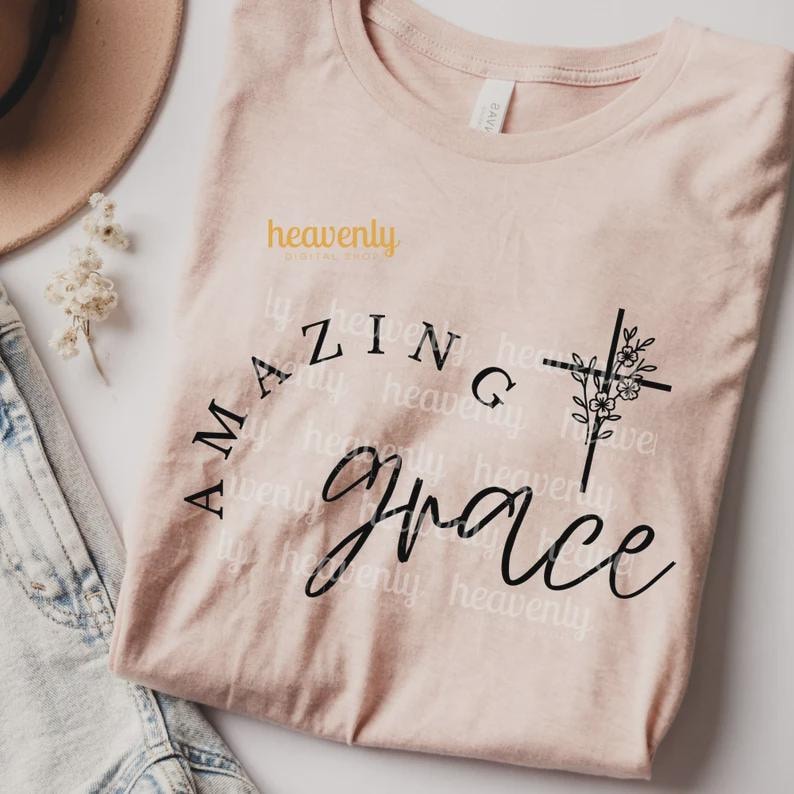 Pu&ograve; includere: Una maglietta rosa chiaro con le parole "Amazing Grace" in corsivo nero. Una croce nera con accenti floreali &egrave; sul lato destro. La maglietta ha anche il testo "heavenly digital shop" in giallo.