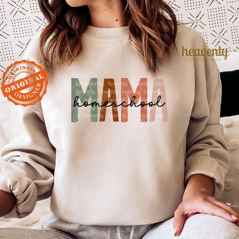 Puede incluir: Una sudadera beige con el texto "MAMA homeschool" en letras coloridas superpuestas. El texto est&aacute; en una fuente estilizada con un aspecto descolorido y vintage.