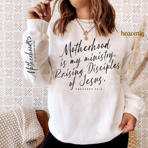 May include: White sweatshirt with black text that reads "Motherhood is my ministry. Raising Disciples of Jesus. Proverbs 22:6"