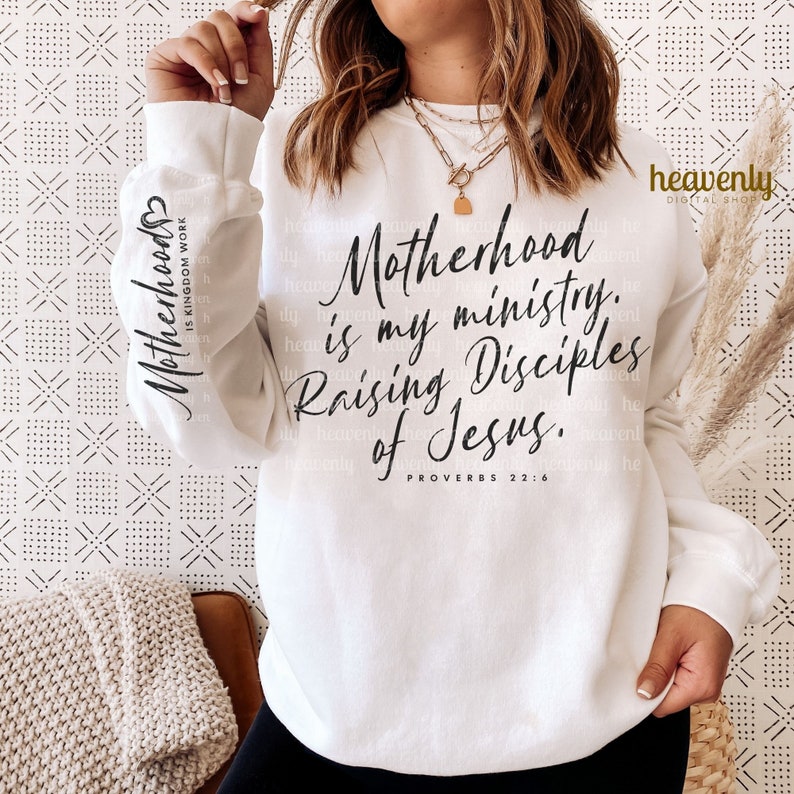 May include: White sweatshirt with black text that reads "Motherhood is my ministry. Raising Disciples of Jesus. Proverbs 22:6"