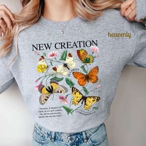 May include: Gray sweatshirt with the text "NEW CREATION" in black lettering. The text is surrounded by a floral design with butterflies in shades of yellow, orange, and brown. The text "IN CHRIST" is written vertically in black lettering. The scripture verse "2 CORINTHIANS 5:17" is printed below the floral design. The text "Therefore, if anyone is in Christ, he is a new creation. The old has passed away: behold, the new has come." is printed below the scripture verse.