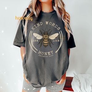 May include: Dark gray t-shirt featuring a circular design with a bee illustration and the text "KIND WORDS ARE LIKE HONEY" with "PROVERBS 16:24" below. The bee is brown and yellow with white wings. The shirt is a short-sleeved, crew neck style.