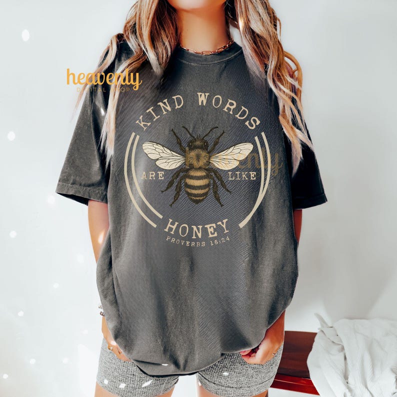 May include: Dark gray t-shirt featuring a circular design with a bee illustration and the text "KIND WORDS ARE LIKE HONEY" with "PROVERBS 16:24" below. The bee is brown and yellow with white wings. The shirt is a short-sleeved, crew neck style.
