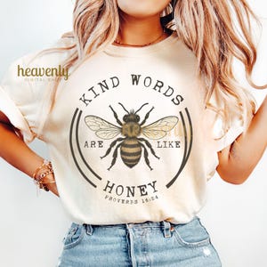 May include: Cream-colored t-shirt featuring a detailed illustration of a bee with the text "Kind Words Are Like Honey" in a circular design. The shirt also includes the Bible verse Proverbs 16:24.