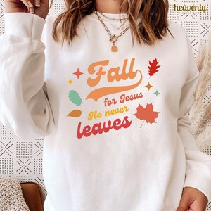 Op de afbeelding: Wit sweatshirt met een kleurrijke afbeelding met de tekst "Fall for Jesus He never leaves" met gestileerde bladeren.