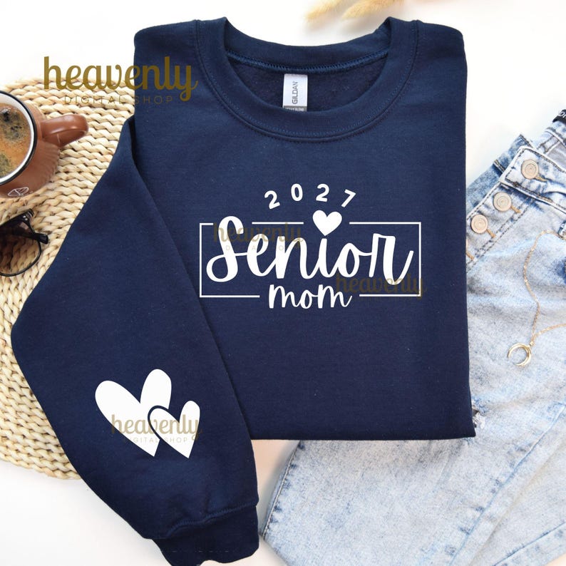 May include: Navy blue sweatshirt with the text "2027 Senior mom" in white. The sweatshirt has two white hearts on the left sleeve. A cup of coffee and sunglasses are visible in the background.