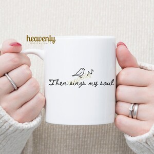 May include: White ceramic mug with the text "Then sings my soul" in a handwritten font, accompanied by a small bird and musical notes. The mug has a handle and is held by a person.