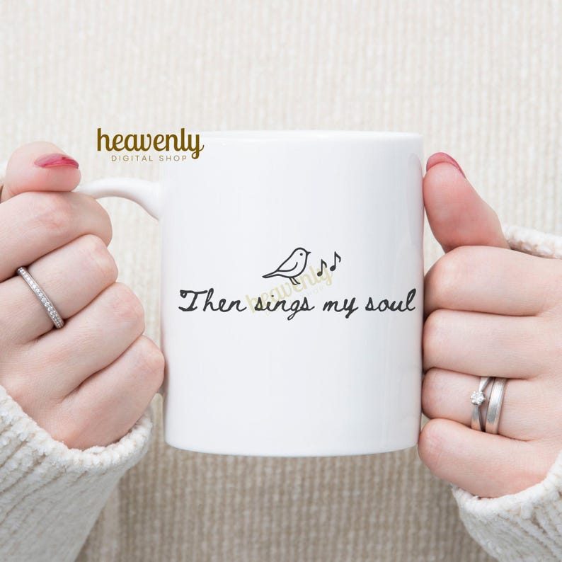 May include: White ceramic mug with the text "Then sings my soul" in a handwritten font, accompanied by a small bird and musical notes. The mug has a handle and is held by a person.
