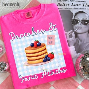 May include: A bright pink t-shirt with the words "Pancakes & Panic Attacks" in white script. The shirt features a graphic of pancakes with blueberries and strawberries. A "Buy 1 Get 1" offer is also displayed.