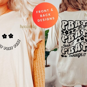 May include: Cream-colored t-shirt with a smiley face design featuring black daisy-shaped eyes and the words "PRAY PRAY PRAY" in a curved font. The back of the shirt has the words "PRAY ON IT, PRAY OVER IT, PRAY THROUGH IT".