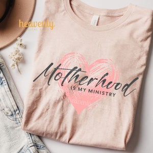 Op de afbeelding: Een roze T-shirt met de tekst "Motherhood is my ministry" in zwarte letters. Een roze hart met een gekrabbelde textuur bevindt zich achter de tekst.