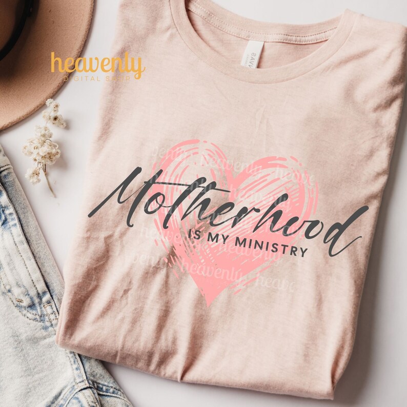 Op de afbeelding: Een roze T-shirt met de tekst "Motherhood is my ministry" in zwarte letters. Een roze hart met een gekrabbelde textuur bevindt zich achter de tekst.