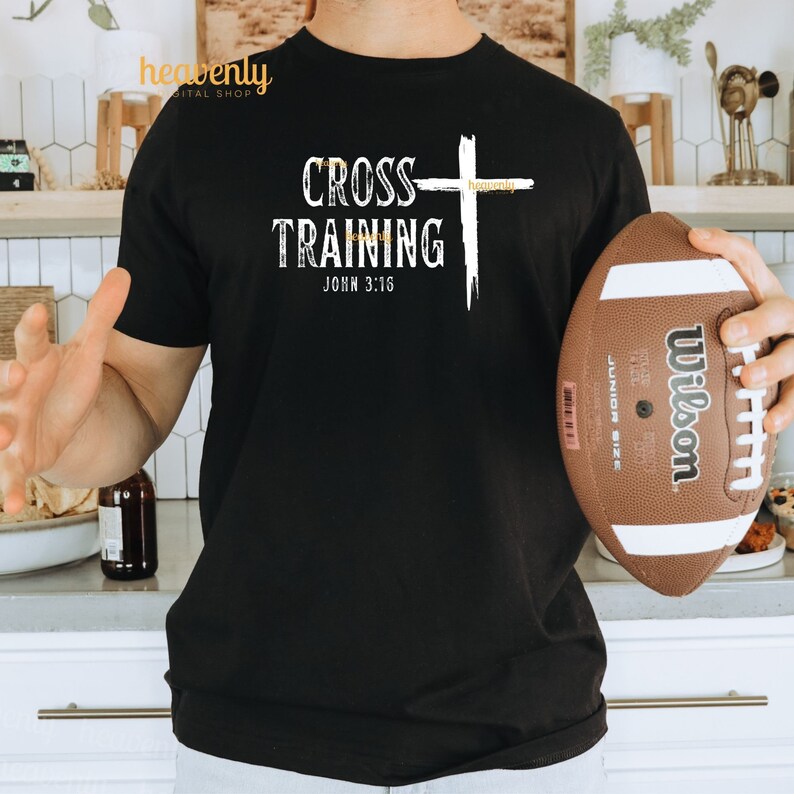 Może przedstawiać: Czarny T-shirt z białym tekstem "Cross Training John 3:16" i białym graficznym krzyżem. Osoba nosi T-shirt i trzyma brązowy futbol.
