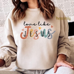 May include: A beige sweatshirt with the words "love like Jesus" in black script and colorful block letters. The word "Jesus" is in orange, pink, teal, and blue. Small hearts are above the words.