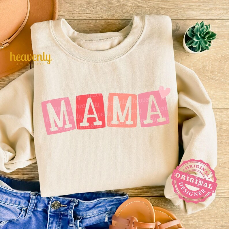 May include: A beige sweatshirt with the word "MAMA" in pink and red block letters with a small pink heart above the word. The sweatshirt is lying on a wooden surface with a pair of blue jeans and sandals.