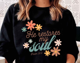 Puede incluir: Sudadera negra con un diseño floral y el texto "He restores my soul" con la referencia bíblica "Salmo 23:3".