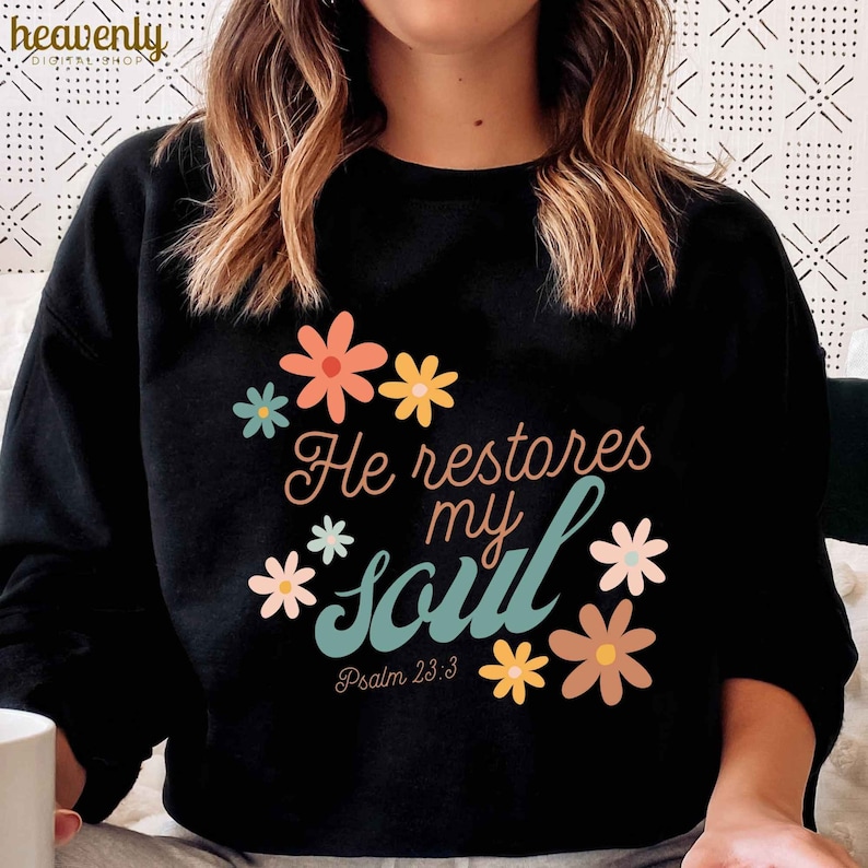 Puede incluir: Sudadera negra con un dise&ntilde;o floral y el texto "He restores my soul" con la referencia b&iacute;blica "Salmo 23:3".