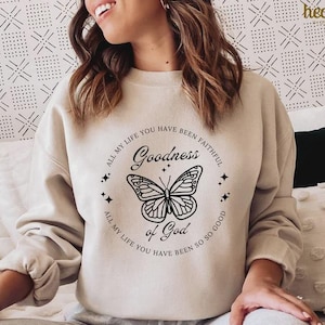 Pu&ograve; includere: Felpa beige con grafica circolare raffigurante una farfalla e le parole "Goodness of God". &Egrave; incluso anche il testo "All my life you have been faithful" e "All my life you have been so so good". La parola "heavenly" si trova nell'angolo in alto a destra.