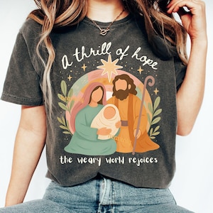 Puede incluir: Camiseta gris oscuro con una ilustración colorida de la escena de la Natividad. El diseño incluye un arcoíris, una estrella y el texto "a thrill of hope" y "the weary world rejoices". La ilustración representa a María, José y el niño Jesús.