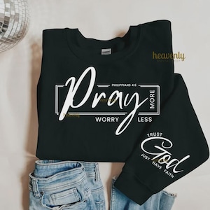 Pu&ograve; includere: Felpa nera con scritta bianca "Pray More, Worry Less" e "Trust God, Just Have Faith". La felpa &egrave; abbinata a jeans blu. Sono visibili anche le parole "heavenly digital shop".