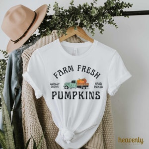 May include: White t-shirt with the text "Farm Fresh Pumpkins" and a green truck graphic carrying pumpkins. The shirt is displayed on a wooden hanger, with a beige hat and denim jacket visible in the background.