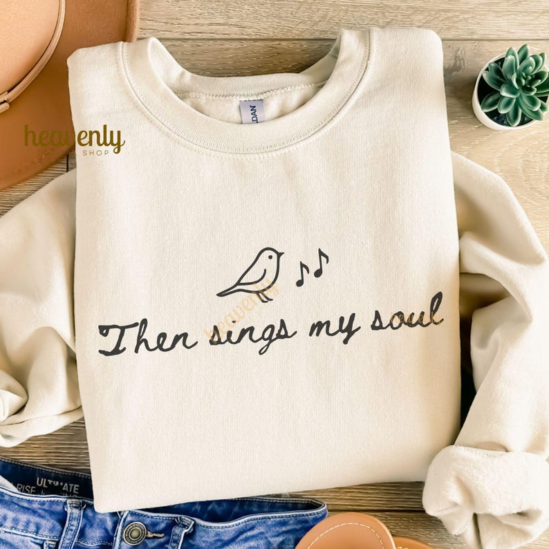 May include: Cream-colored sweatshirt with a crew neck. The front features a black bird illustration with musical notes and the text "Then sings my soul" in a handwritten style. The sweatshirt is folded on a wooden surface.