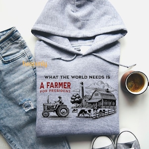 May include: Gray hooded sweatshirt with the text "What the world needs is a farmer for president" printed on the front. The design features a cartoon illustration of a farmer driving a tractor in front of a farm.