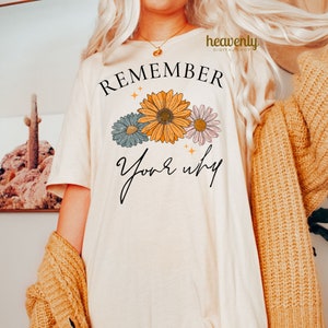 K&ouml;nnte beinhalten: Ein wei&szlig;es T-Shirt mit dem Text "REMEMBER your why" in schwarzer Schrift. Der Text ist von drei stilisierten Blumen in Blau-, Gelb- und Rosat&ouml;nen umgeben.